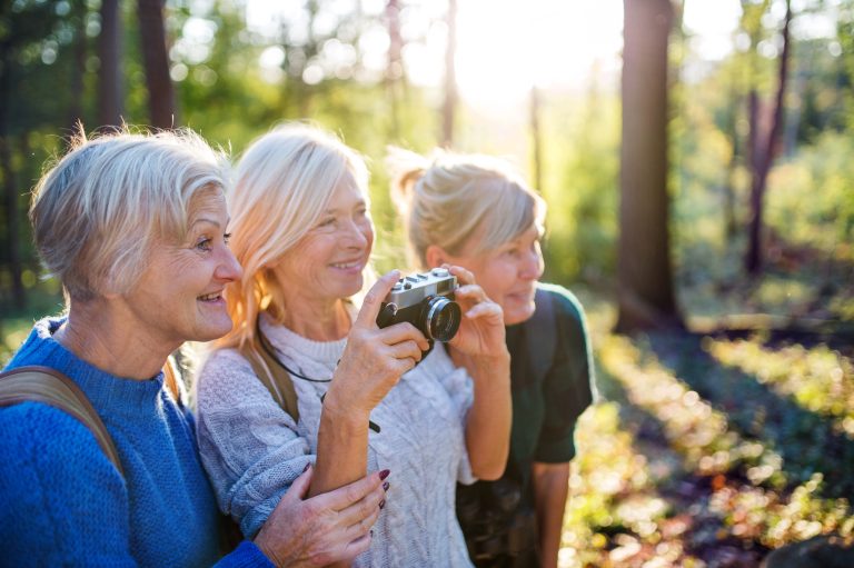 Mature female friends walking outdoors in a wooded area, taking photos with a camera.