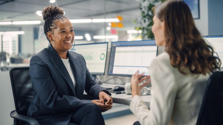 Two female colleagues talking in front of computer screens.